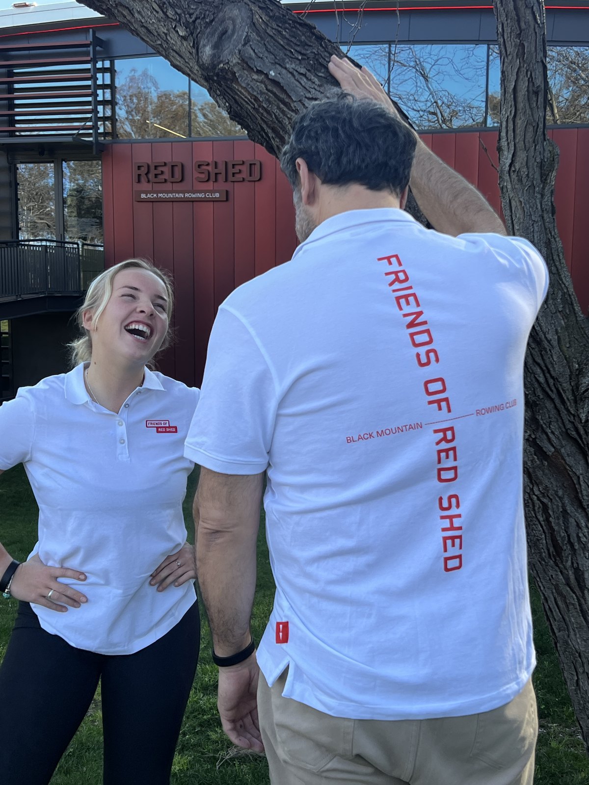 Two people in white Friends of Red Shed polos outside the Red Shed building, the back print reading FRIENDS OF RED SHED above Black Mountain Rowing Club