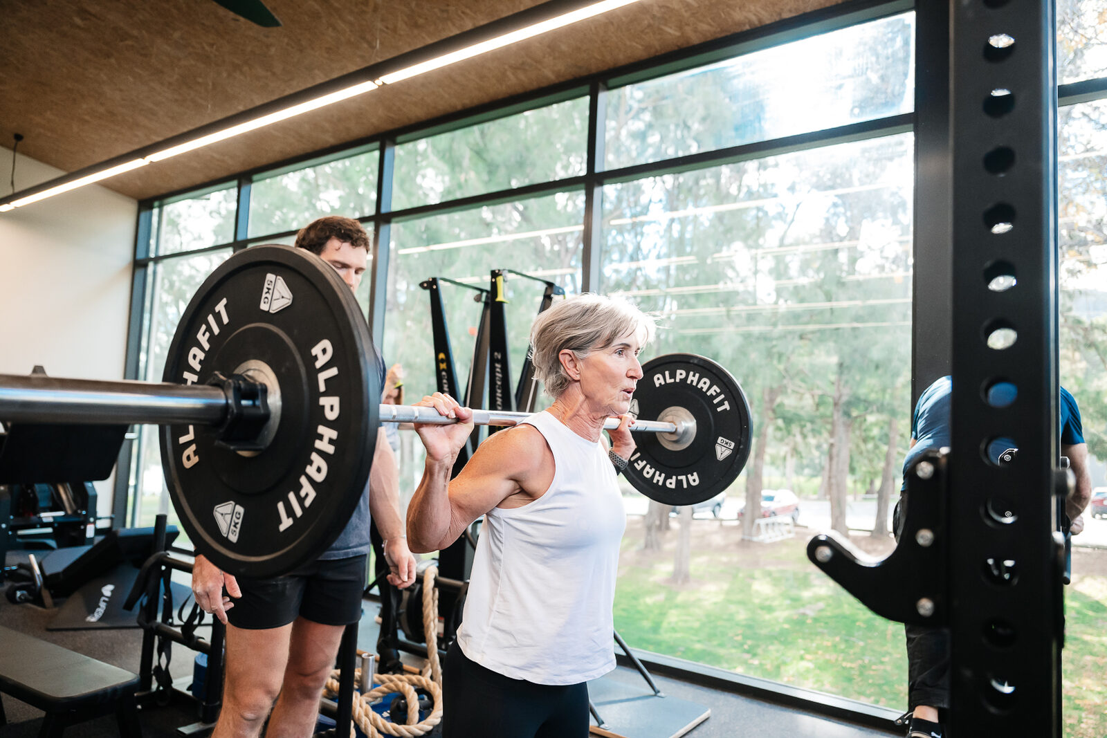 Red Shedder squatting a loaded barbell in the rack at RSX Strength Club, coach standing behind her