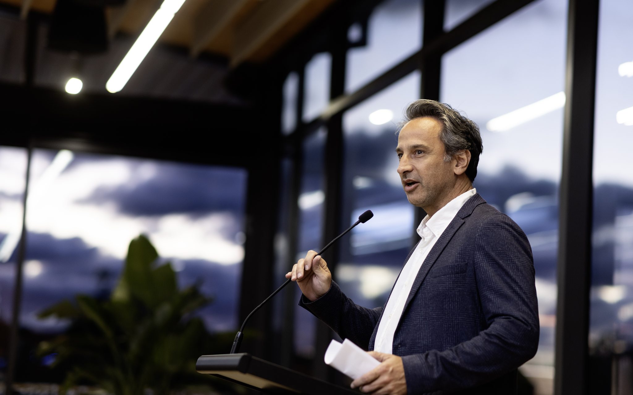 A speaker at the lectern in the Telopea Room at dusk, the lake behind the glass